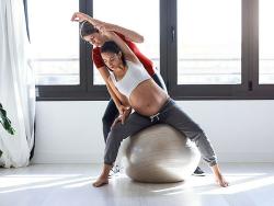 Eine junge schwangere Frau balanaciert auf einem Sitzball und erhält ein Stretching von einer Therapeutin